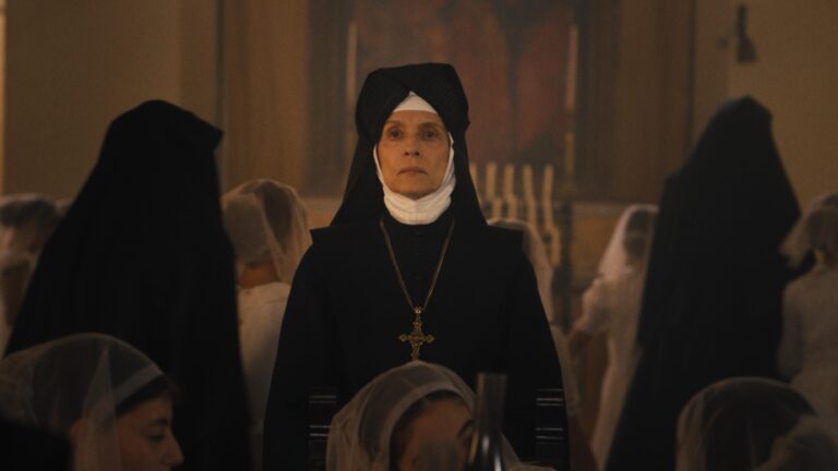 Kadr z filmu Omen: Początek. Starsza zakonnica w tradycyjnym czarnym habitu i z welonem stojąca na tle innych zakonnic w kościele.