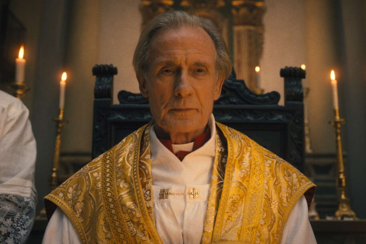 Kadr z filmu Omen: Początek. Starszy mężczyzna w stroju duchownym stoi przed ołtarzem z zapalonymi świecami.