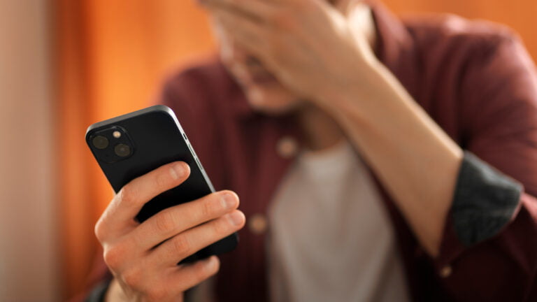 Osoba trzymająca smartfon i zakrywająca twarz ręką w geście zmartwienia lub zmęczenia.