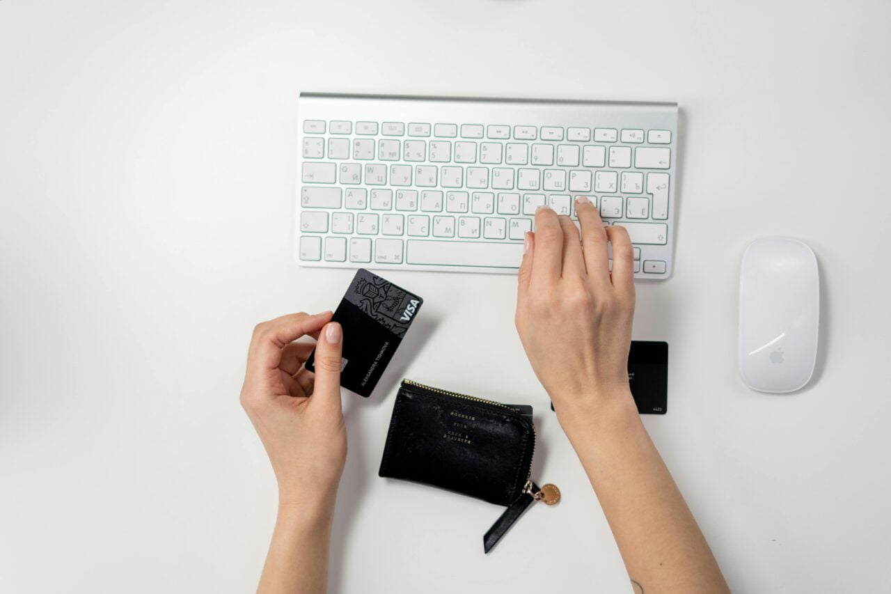 Dłonie osoby piszącej na białej klawiaturze komputera z leżącą obok białą myszką, trzymającej w jednej ręce czarną kartę kredytową, z czarnym portfelem na biurku.