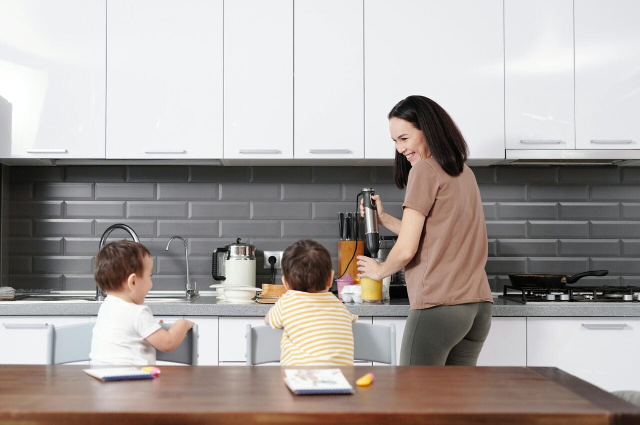 Kobieta przygotowuje napój w blenderze w nowoczesnej kuchni, dwójka małych dzieci siedzi przy stole kuchennym.