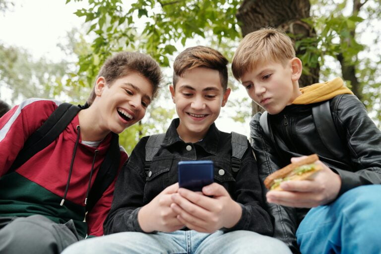 Trzej uśmiechnięci nastolatkowie siedzący na zewnątrz i patrzący na telefon komórkowy, który trzyma jeden z nich.