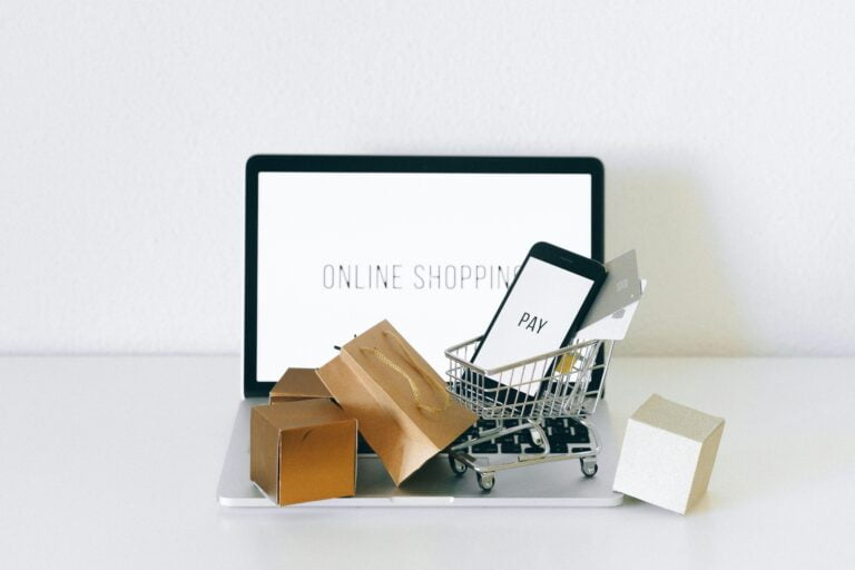 Kompozycja przedstawiająca koncept zakupów online z tabletem wyświetlającym napis "ONLINE SHOPPING", małym wózkiem na zakupy z telefonem komórkowym i kartami kredytowymi oraz kilkoma pudełkami na laptopie i obok, symbolizując zamówienia internetowe.