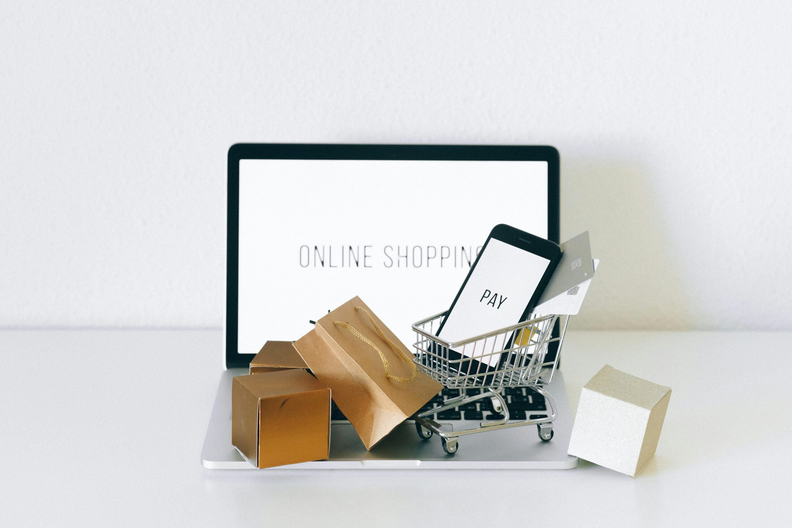 Kompozycja przedstawiająca koncept zakupów online z tabletem wyświetlającym napis "ONLINE SHOPPING", małym wózkiem na zakupy z telefonem komórkowym i kartami kredytowymi oraz kilkoma pudełkami na laptopie i obok, symbolizując zamówienia internetowe.