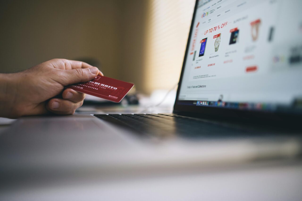 Osoba trzymająca czerwoną kartę kredytową przy laptopie z otwartą stroną sklepu internetowego.