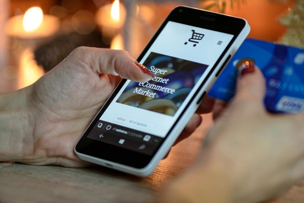 Osoba przeglądająca stronę sklepu internetowego na smartfonie, trzymając w drugiej ręce kartę kredytową, z rozmytymi światłami w tle.