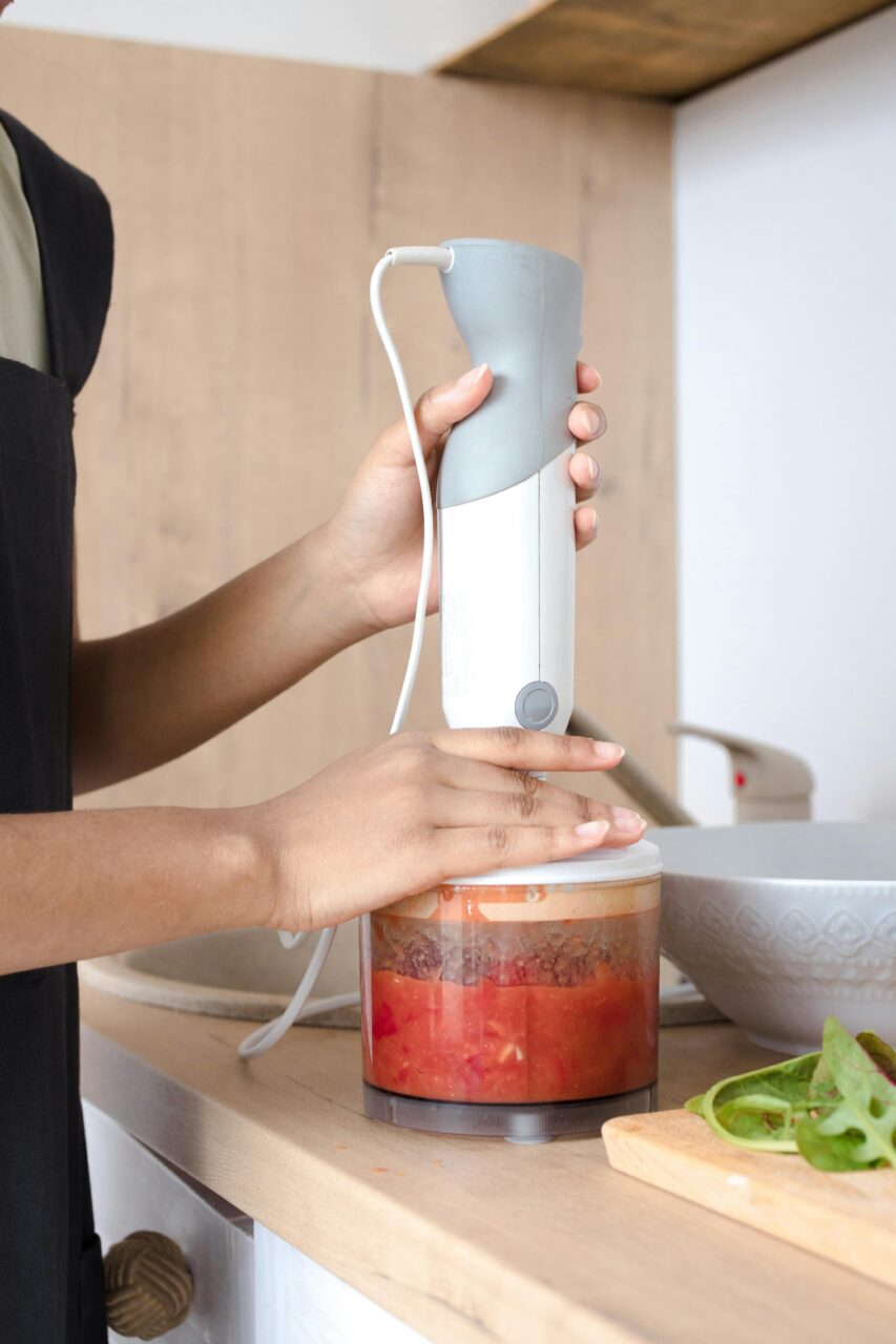 Blender ręczny, osoba używa ręcznego blendera do miksowania składników w przezroczystym pojemniku na drewnianym blacie kuchennym.