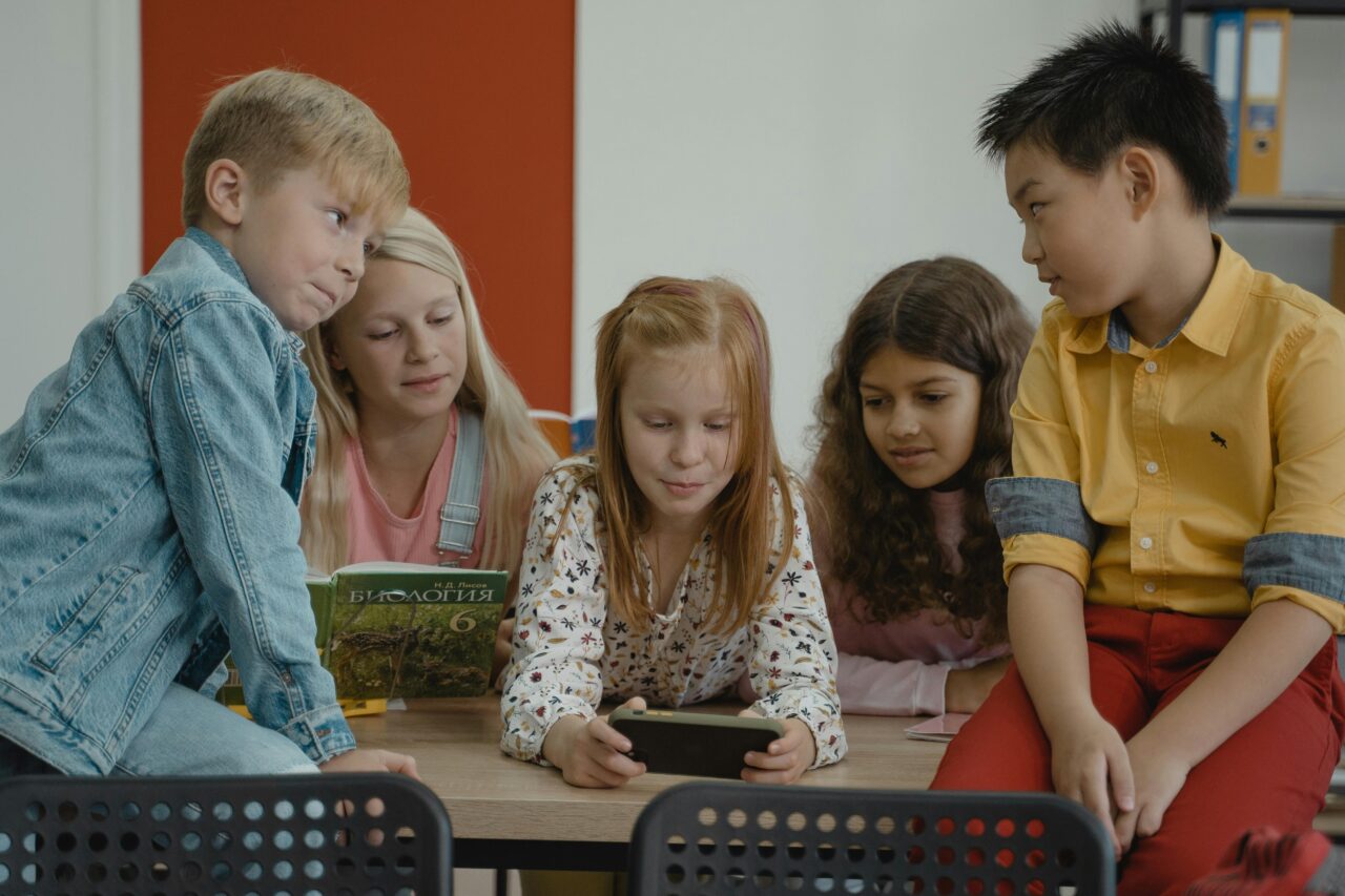 Grupa dzieci w różnym wieku siedzi wokół stołu w klasie i wspólnie patrzy na ekran tabletu trzymanego przez dziewczynkę z rudymi włosami.