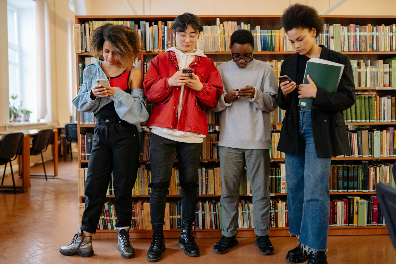Czterech młodych ludzi stoi obok siebie, skupionych na używaniu swoich smartfonów w bibliotece z półkami pełnymi książek w tle.