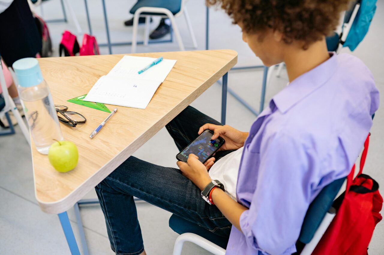 Uczeń siedzi przy ławce szkolnej i gra na smartfonie, na biurku leży zeszyt, długopis, linijka, okulary i zielone jabłko obok butelki z wodą.