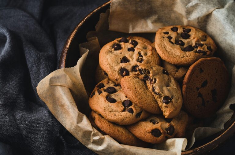 Ciasteczka z kawałkami czekolady symbolizujące pliki cookie w ceramicznym naczyniu na papierze do pieczenia, na tle ciemnej tkaniny.