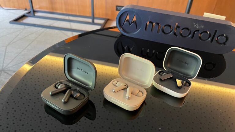 Dwa otwarte etui na słuchawki bezprzewodowe marki Motorola, z widocznymi na pierwszym planie słuchawkami, na szklanym stoliku odbijającym ich obraz, w tle logo Motorola.