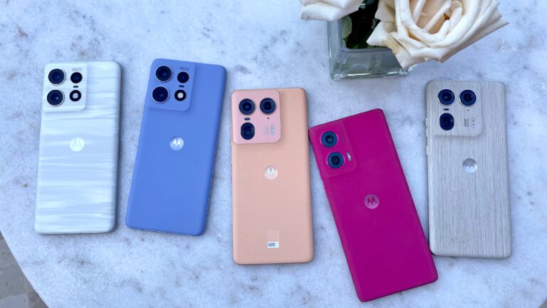 Pięć smartfonów różnych kolorów z aparatami rozmieszczonymi w kwadrat na marmurowym tle obok szklanego wazonu z białą różą.