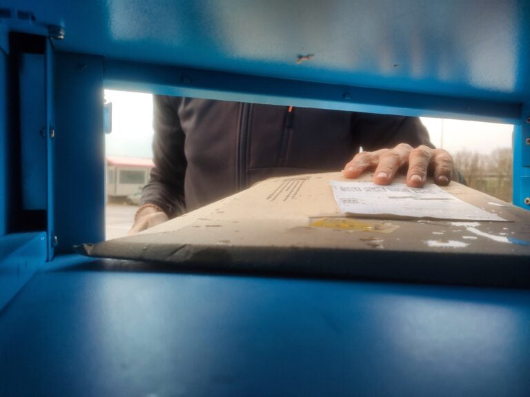 widok ze skrytki automatu paczkowego, do którego wkładana jest płaska przesyłka w kartonie z naklejoną etykietą przez mężczyznę w brązowej bluzie