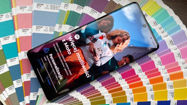 Smartfon leżący na próbniku kolorów Pantone, wyświetlający grafikę reklamową z osobami.