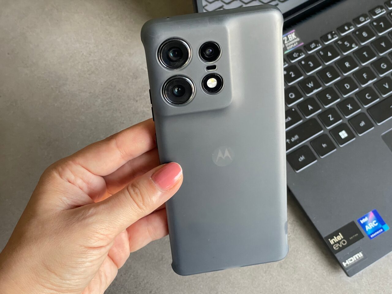 Ręka trzymająca smartfon marki Motorola z potrójnym aparatem, na tle klawiatury laptopa.