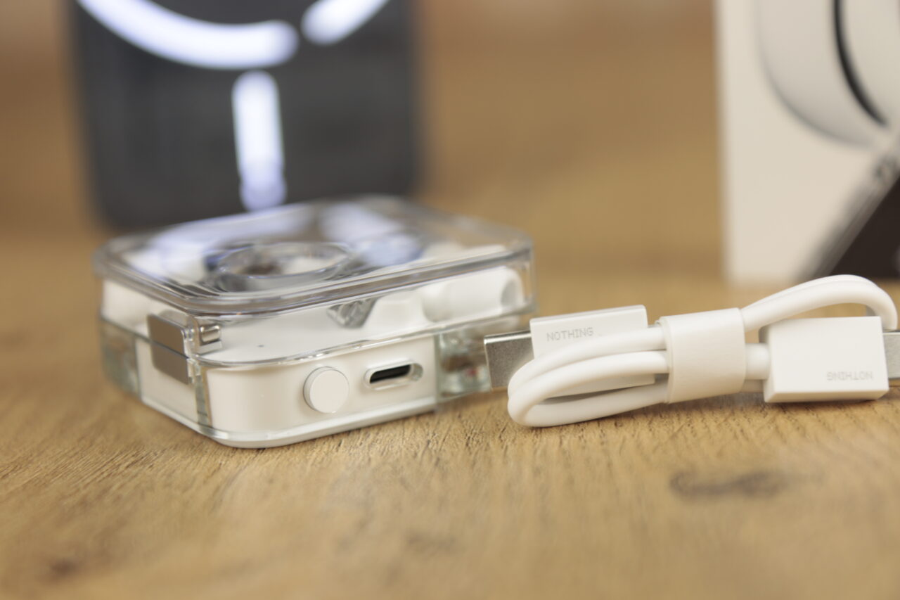 Słuchawki Nothing Ear w przezroczystym etui z białym kablem USB na drewnianym stole, w tle opakowanie produktu.