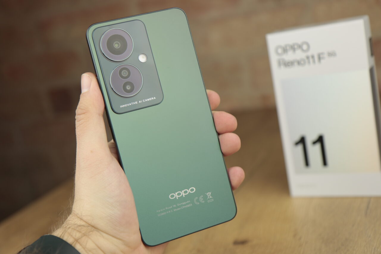 Tylna część zielonego smartfona Oppo trzymanego w dłoni z widocznym zespołem trzech aparatów i inskrypcją "Innovative AI Camera", w tle pudełko z napisem "Oppo Reno11 F".