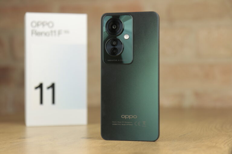 Smartfon OPPO Reno11 F 5G koloru zielonego z potrójnym aparatem i napisem "INNOVATIVE AI CAMERA" na tylnej obudowie, ustawiony na drewnianym blacie obok pudełka z wyraźnie widoczną cyfrą "11".