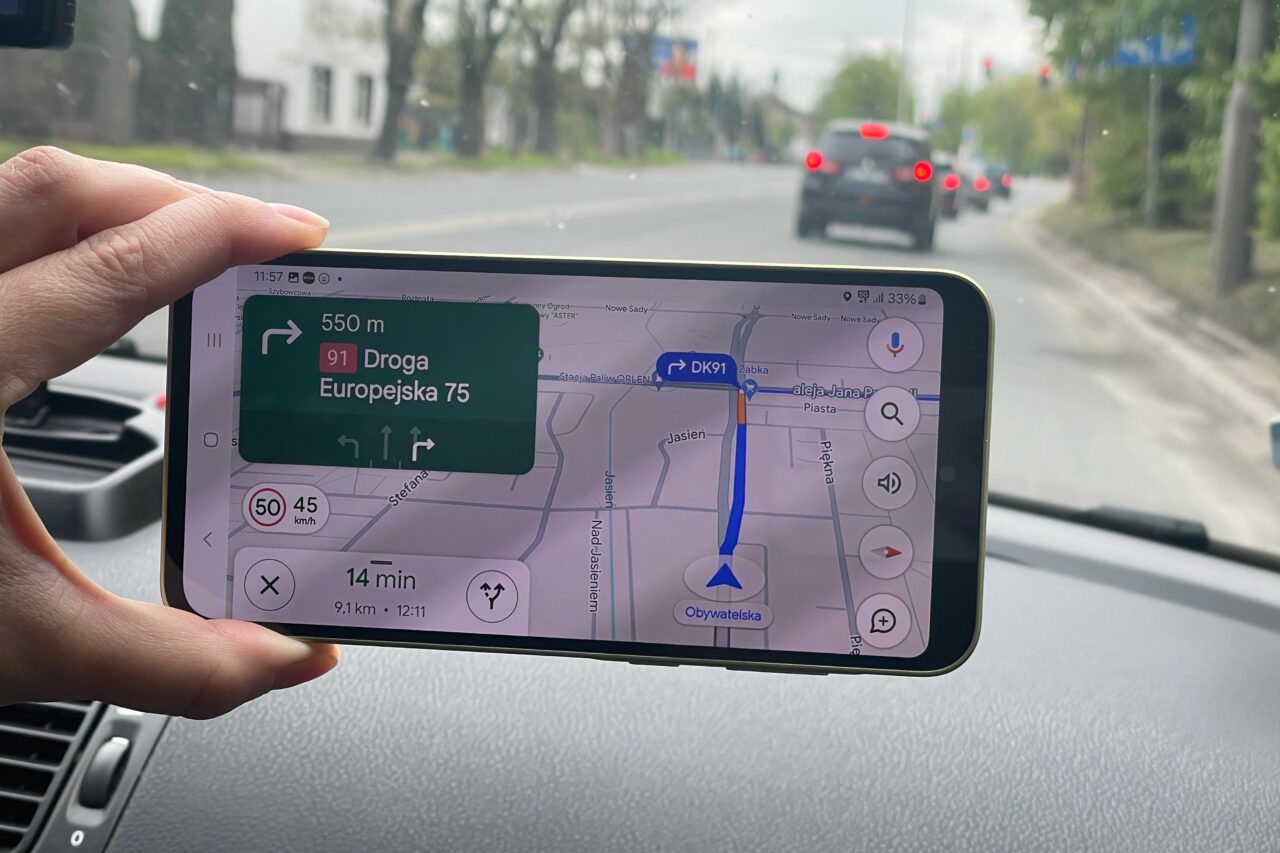 Ręka trzymająca smartfon z włączoną nawigacją GPS w samochodzie, pokazującą trasę na mapie.