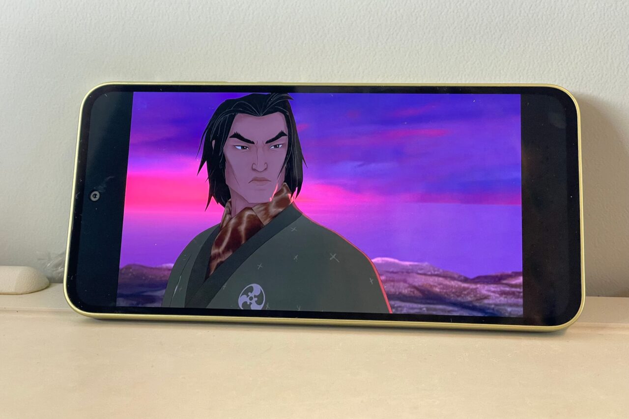 Animowana postać mężczyzny o azjatyckich rysach na ekranie telefonu, na tle zachodzącego słońca i gór.