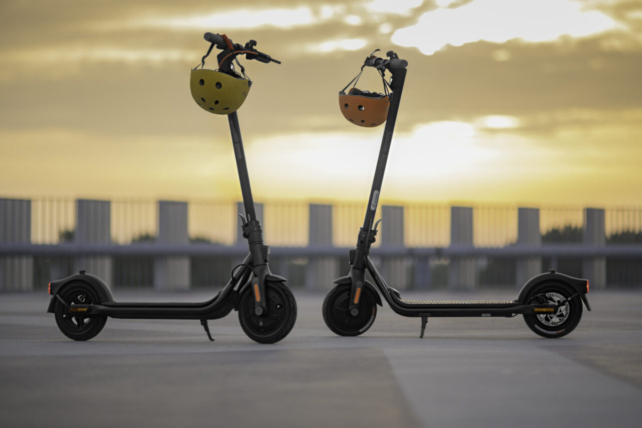 Dwa elektryczne hulajnogi ze skuterami na parkingu z kaskami z wizerunkiem owoców na kierownicach, zachód słońca w tle.