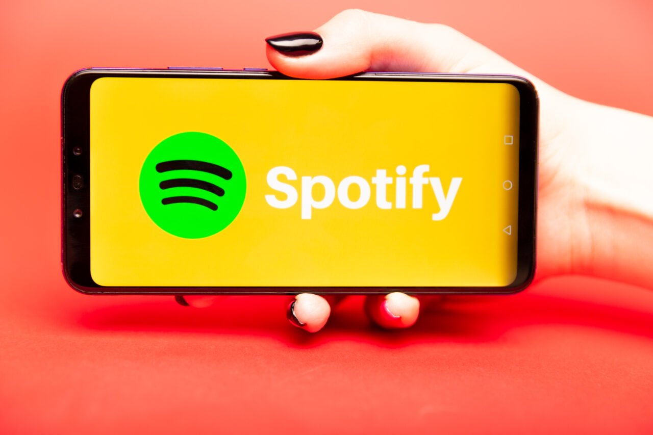 Dłoń trzymająca smartfon z aplikacją Spotify na żółtym tle ekranie.