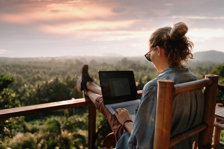 Kobieta praca zdalna na laptopie na zewnątrz na drewnianym tarasie z widokiem na zalesiony krajobraz przy zachodzie słońca.