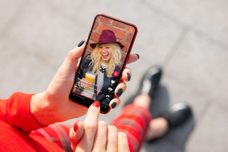 Osoba ogląda wideo na smartfonie przedstawiające uśmiechniętą kobietę w kapeluszu pijącą sok.