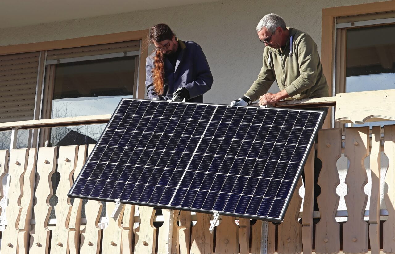 Dwóch mężczyzn montuje panel słoneczny na drewnianym balkonie. Fotowoltaika to wspólna sprawa.