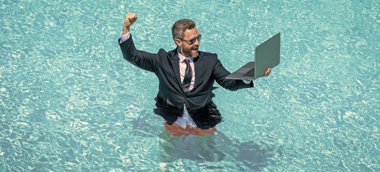 Mężczyzna w garniturze i okularach przeciwsłonecznych unosi rękę w geście triumfu, stojąc w wodzie basenu i trzymając laptopa.