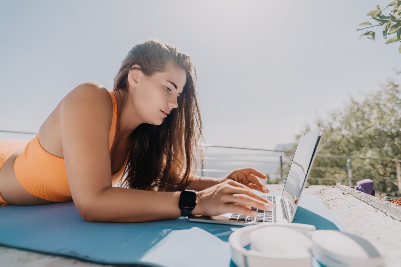 Kobieta w pomarańczowym sportowym topie korzysta z laptopa na zewnątrz na jasnoniebieskiej macie do jogi, w tle widoczne drzewa i jezioro.