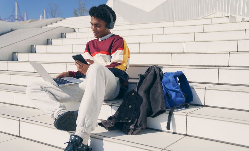 Młody mężczyzna z słuchawkami Sony na uszach siedzi na zewnątrz na schodach i używa laptopa trzymając telefon komórkowy w dłoni, obok niego leży plecak i kurtka.
