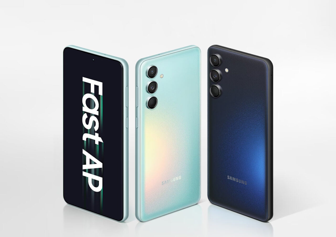 Trzy smartfony Samsung Galaxy M55 5G stojące pionowo z wyświetlaczami zwróconymi w stronę widza; jeden w kolorze czarnym z napisem "FastAP" na ekranie, drugi w kolorze pastelowym, trzeci w kolorze granatowym. Wszystkie posiadają potrójny układ aparatów umieszczony wzdłuż krawędzi tyłu obudowy.