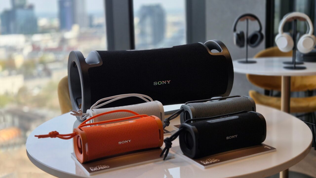 Różne modele przenośnych głośników marki Sony ułożone na biurku z widokiem na miasto przez okno w tle.
