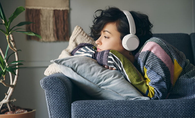 Osoba odpoczywająca na kanapie z nałożonymi słuchawkami na uszach i wtulona w kolorowe poduszki. Kupiła świetne słuchawki Sony na promocji.