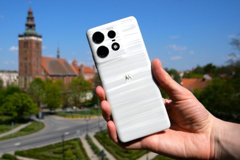 Dłoń trzymająca biały smartfon z potrójnym aparatem na tle miasta.