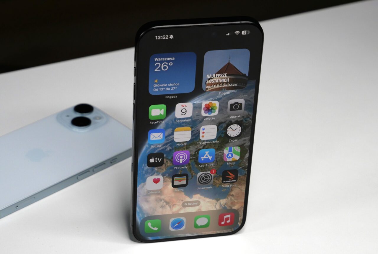 Atak na iPhone'y. Smartfon z włączonym ekranem pokazującym interfejs głównego ekranu z aplikacjami, umieszczony na białym tle z innym smartfonem z tyłu.