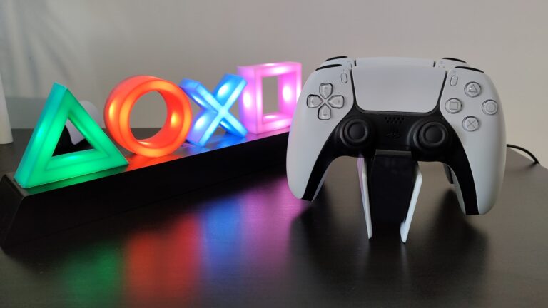 Podświetlane symbole konsoli PlayStation (trójkąt, koło, krzyżyk i kwadrat) w różnych kolorach z kontrolerem do gier na pierwszym planie, umieszczonym na stojaku. Stacja dokująca ładuje kontrolery do PS5