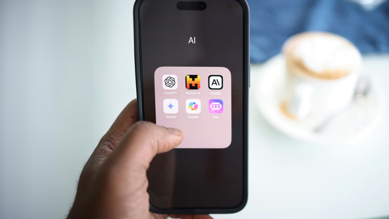 Apple iPhone wyświetlający folder z aplikacjami używającymi sztucznej inteligencji. W tle rozmazana filiżanka z kawą.