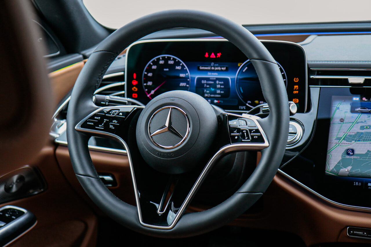 Wnętrze Mercedesa klasy E podczas testu, z widokiem na kierownicę i panel z ekranami.