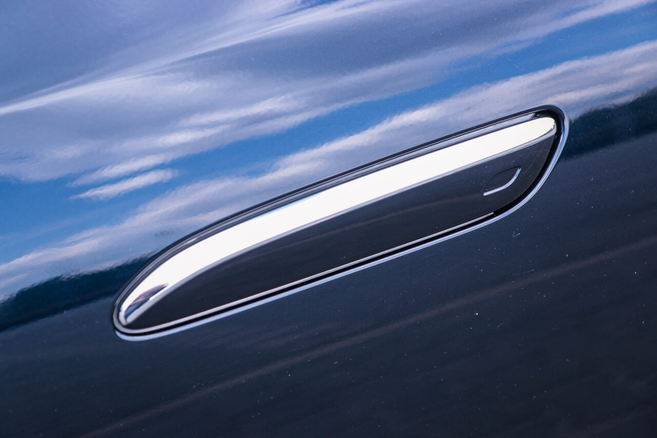 Detal testu Mercedes Klasy E: zbliżenie na chromowaną klamkę samochodową z odbiciem chmur.