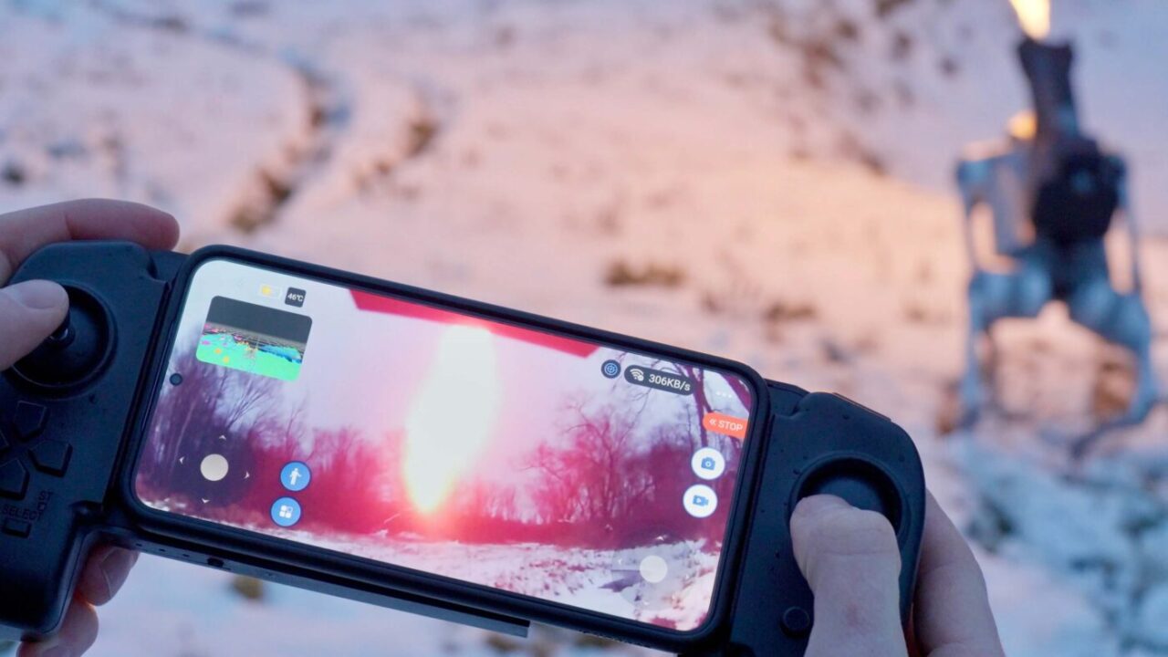 Ręka trzymająca kontroler do smartfona, który wyświetla eksplozję na śnieżnym tle, w tle nieostra postać człowieka.