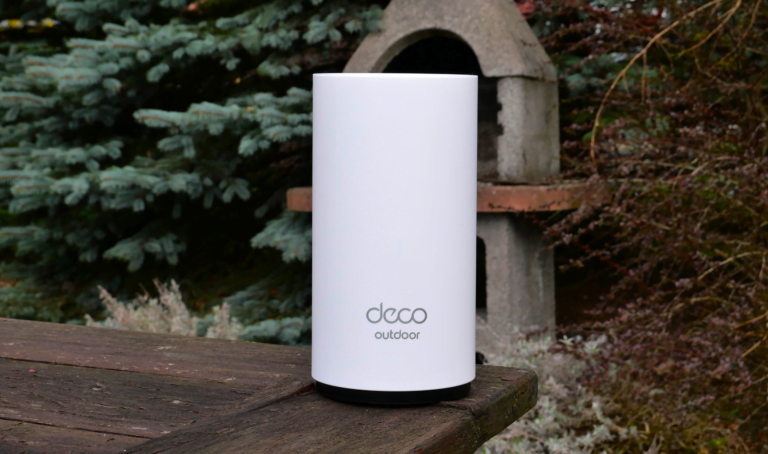 Biały, cylindryczny router WiFi z napisem "deco outdoor" na pierwszym planie, ustawiony na drewnianej poręczy z roślinnością i kamiennym słupem w tle.
