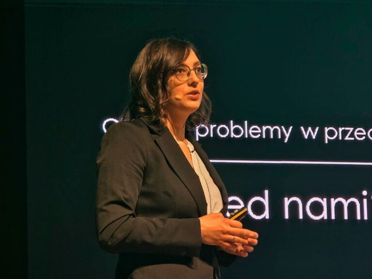 Magdalena Wilczyńska, szefowa POIC NASK. Kobieta w okularach i czarnym garniturze prezentuje wykład, stojąc przed ekranem z białym tekstem.