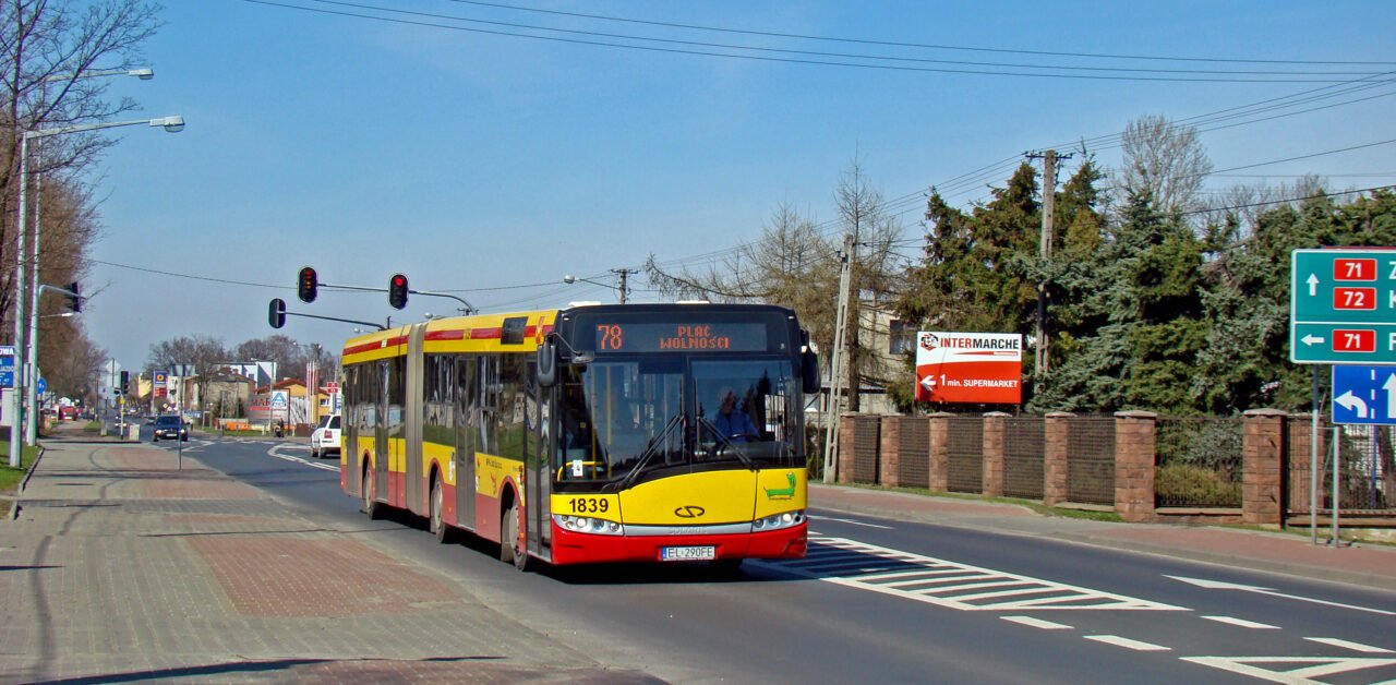 Czerwono-żółty autobus na ulicy miejskiej, sygnalizacja świetlna na czerwono, tablice drogowe i reklama supermarketu Intermarché.