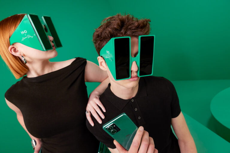 Dwóch młodych dorosłych w czarnych ubraniach, noszących zielone, geometryczne okulary przeciwsłoneczne w otoczeniu zielonego tła.