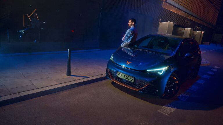 Mężczyzna stoi obok nowoczesnego elektrycznego samochodu na oświetlonym parkingu w nocy.