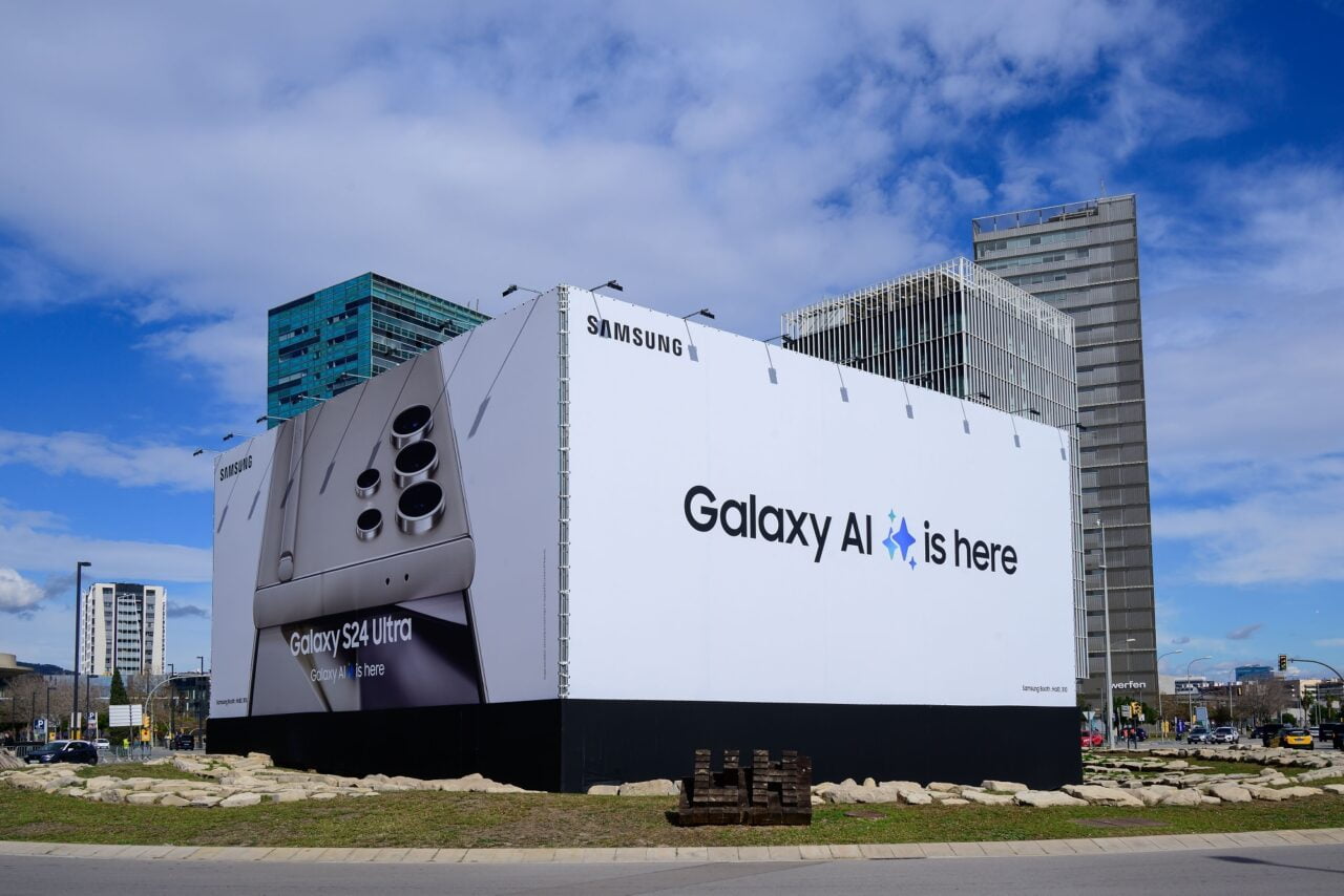 Reklama na dużym billboardzie prezentująca Samsung Galaxy S24 Ultra z napisem „Galaxy AI is here” w tle miejskiego krajobrazu z biurowcami.