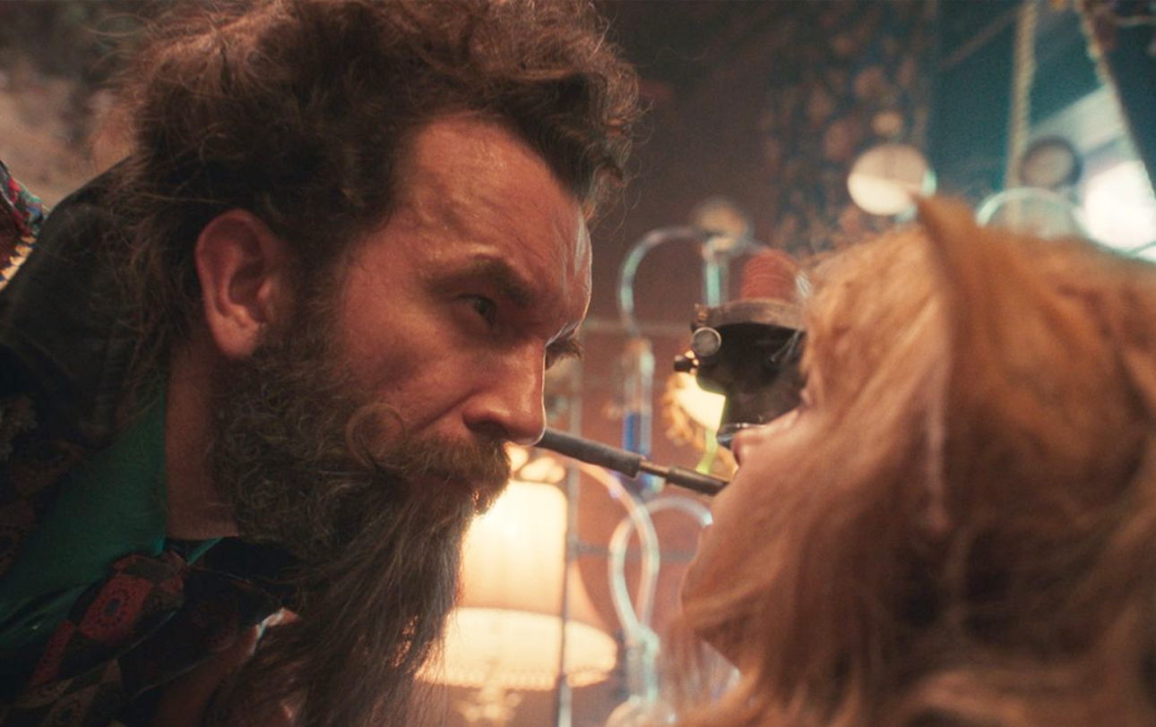 Kadr z filmu Akademia Pana Kleksa. Zbliżenie twarzy mężczyzny z bujną brodą patrzącego na kobietę z długimi rudymi włosami za pomocą specjalnego przyrządu optycznego.